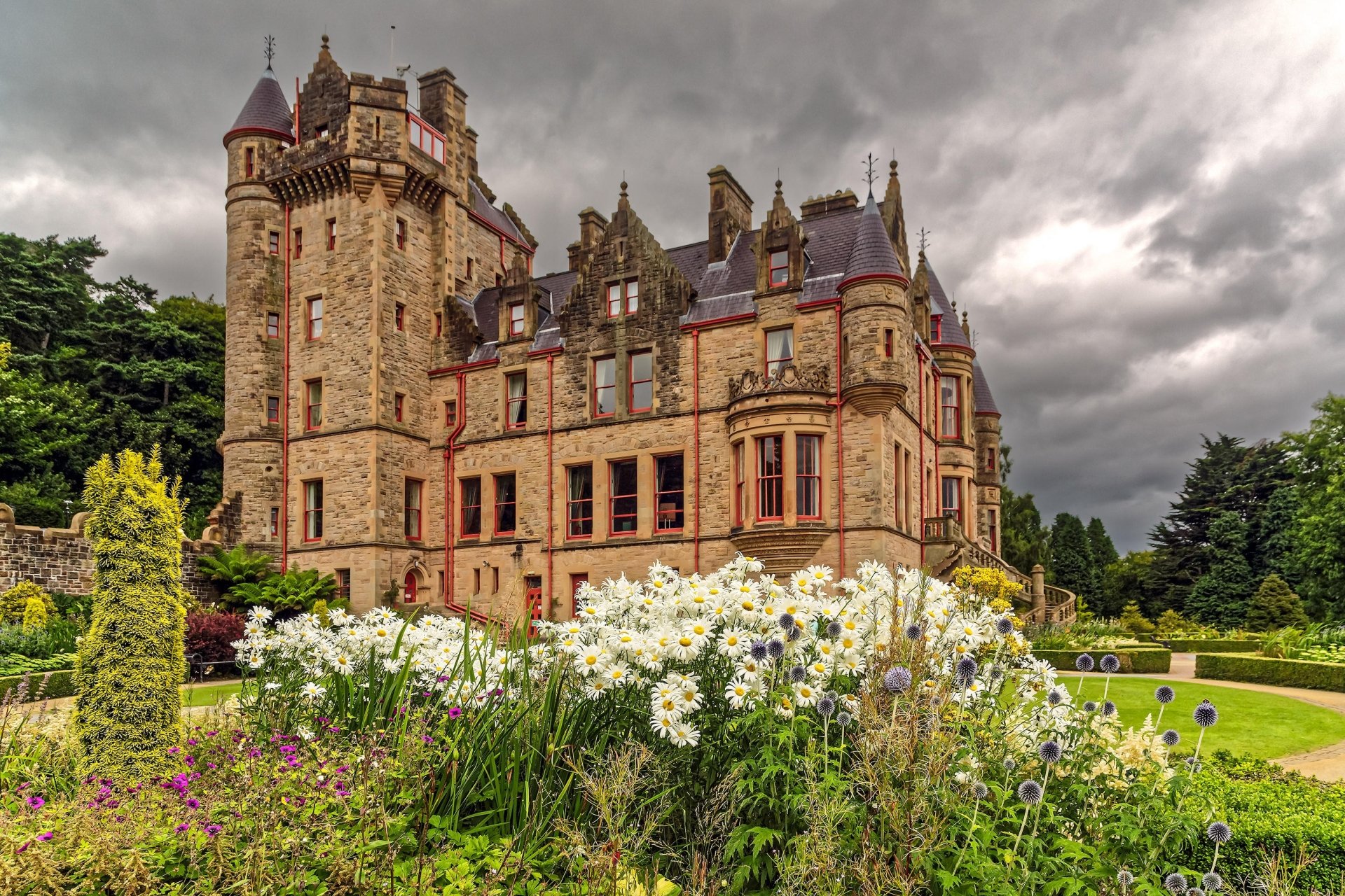 Belfast Castle in Ireland stands majestically amid lush gardens with blooming flowers under a dramatic cloudy sky, showcasing historic architecture.