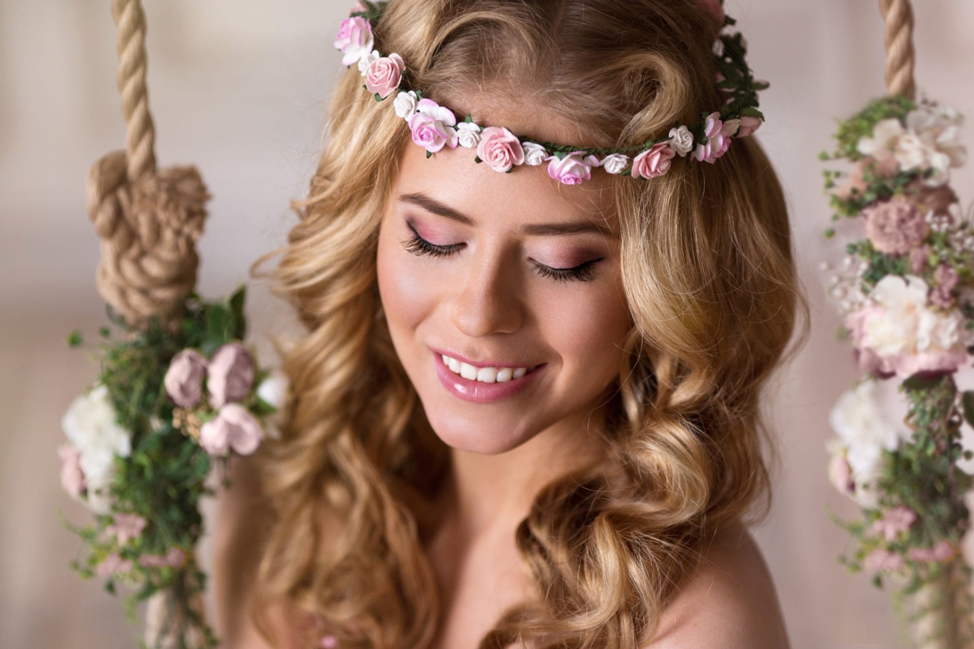 Portrait of a smiling woman with makeup and a floral wreath in her wavy hair, surrounded by soft flowers, creating a serene and radiant mood.