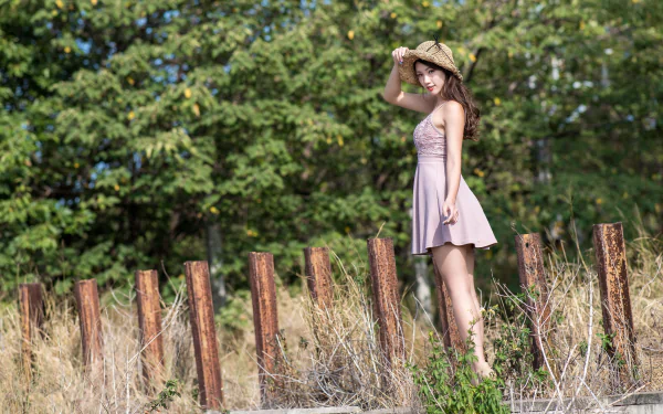 Brunette woman in a pink dress and straw hat poses by wooden posts in a sunlit grassy field — 4K Ultra HD PC desktop wallpaper.