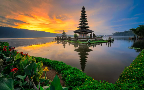 Sunset over Pura Ulun Danu Bratan temple in Bali, Indonesia, reflecting in calm water surrounded by lush greenery and flowers, showcasing serene natural beauty.