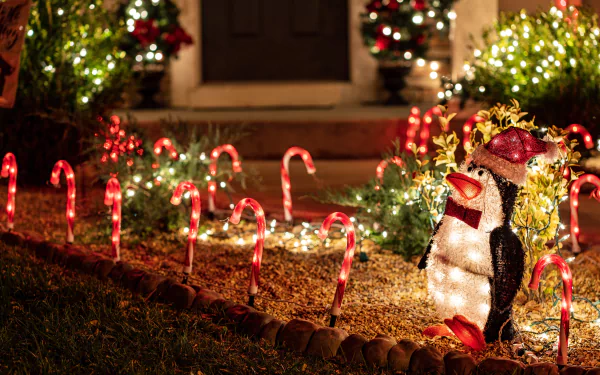 8K Ultra HD PC desktop wallpaper/background of a lit penguin lawn ornament amid candy-cane lights, Christmas ornaments and warm holiday porch lights.