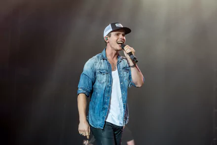 HD wallpaper featuring a performer on stage, wearing a baseball cap and denim jacket, energizing the crowd with a microphone in hand.