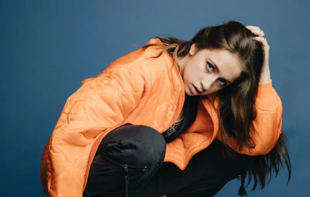 HD desktop wallpaper of Tate McRae posing in an orange jacket against a blue background.