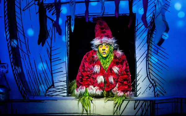 HD wallpaper of The Grinch in red costume peeking out from a snowy window, from Dr. Seuss' The Grinch Musical.