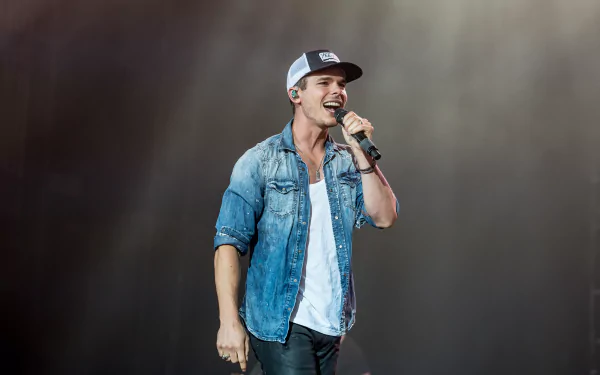 HD wallpaper featuring a performer on stage, wearing a baseball cap and denim jacket, energizing the crowd with a microphone in hand.