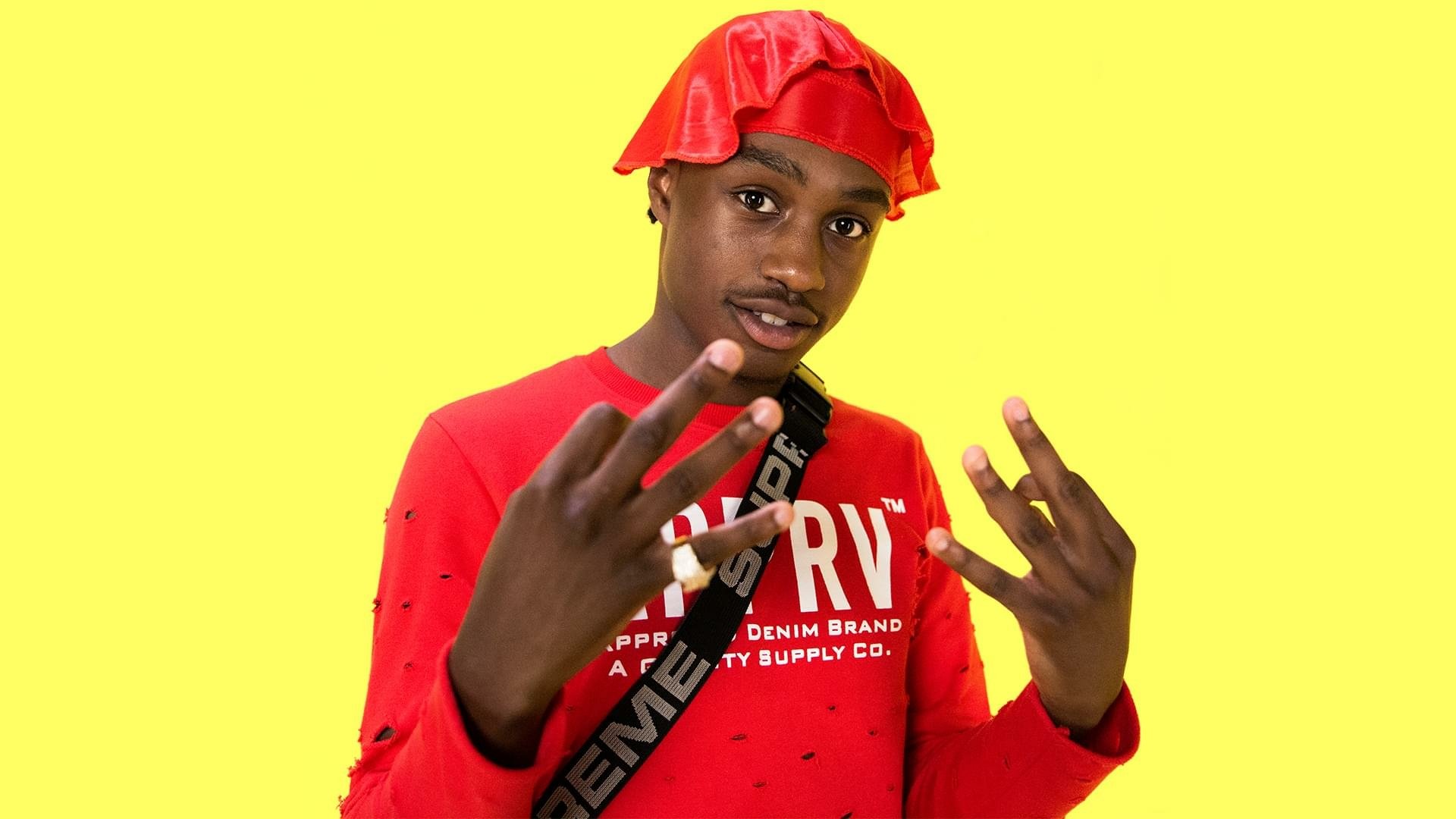 HD desktop wallpaper featuring a person in a red outfit and bucket hat against a yellow background, gesturing with their hands.