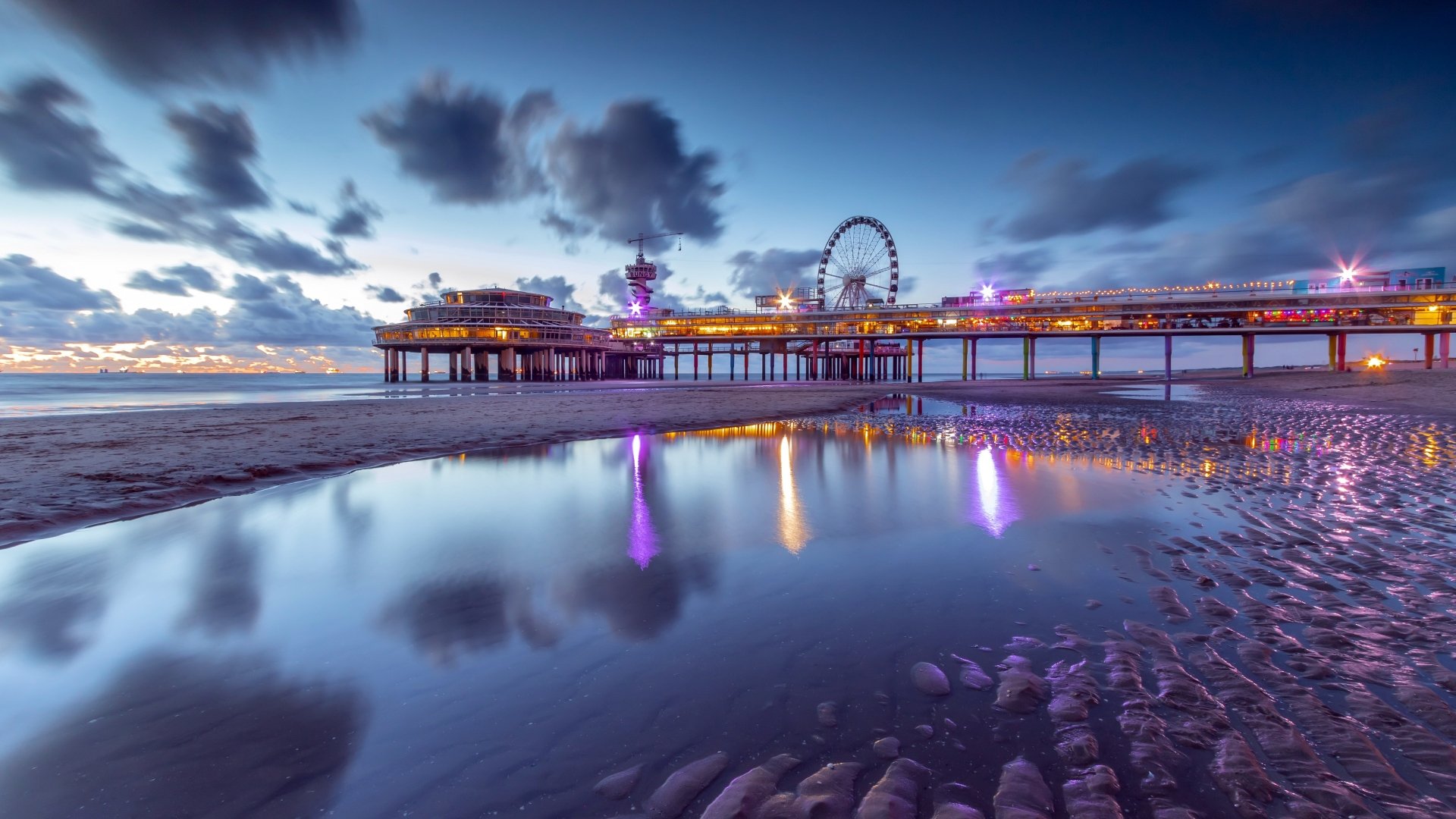 4K Ultra HD PC wallpaper of Scheveningen at night: a man-made pier and Ferris wheel on the horizon, ocean and wet sand reflecting colorful lights.