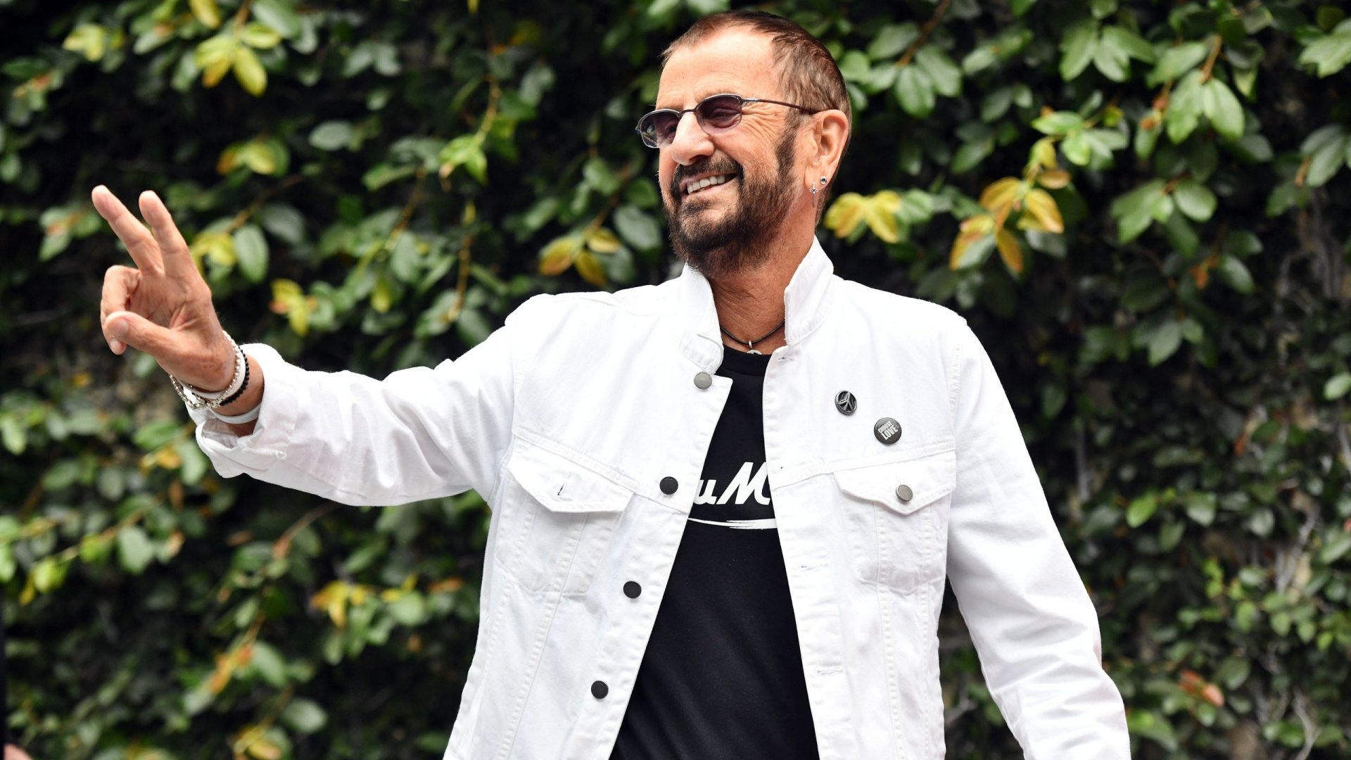 HD desktop wallpaper featuring a cheerful man flashing a peace sign with a green leafy background.
