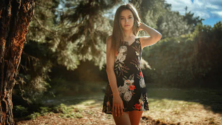 Brunette woman model in a floral dress stands in a sunlit outdoor setting with blurred trees creating a depth of field effect, captured in HD quality for a desktop wallpaper.
