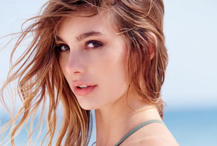 Close-up portrait of a red-haired female model with windblown hair and subtle makeup against a pale blue seaside background — 2K Quad HD PC desktop wallpaper.