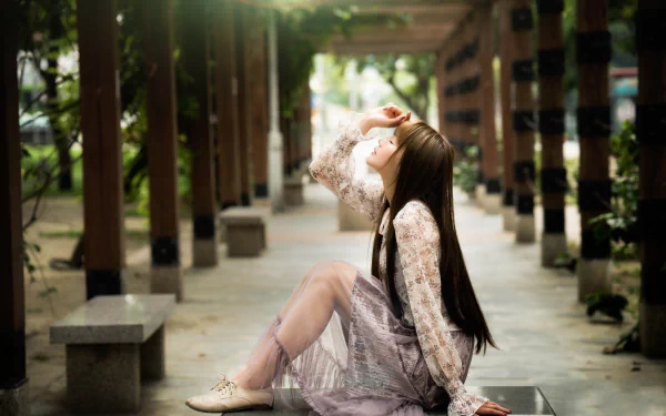 An Asian brunette woman with long hair sits gracefully on a bench in a softly lit outdoor corridor, captured with a shallow depth of field in 8K Ultra HD.