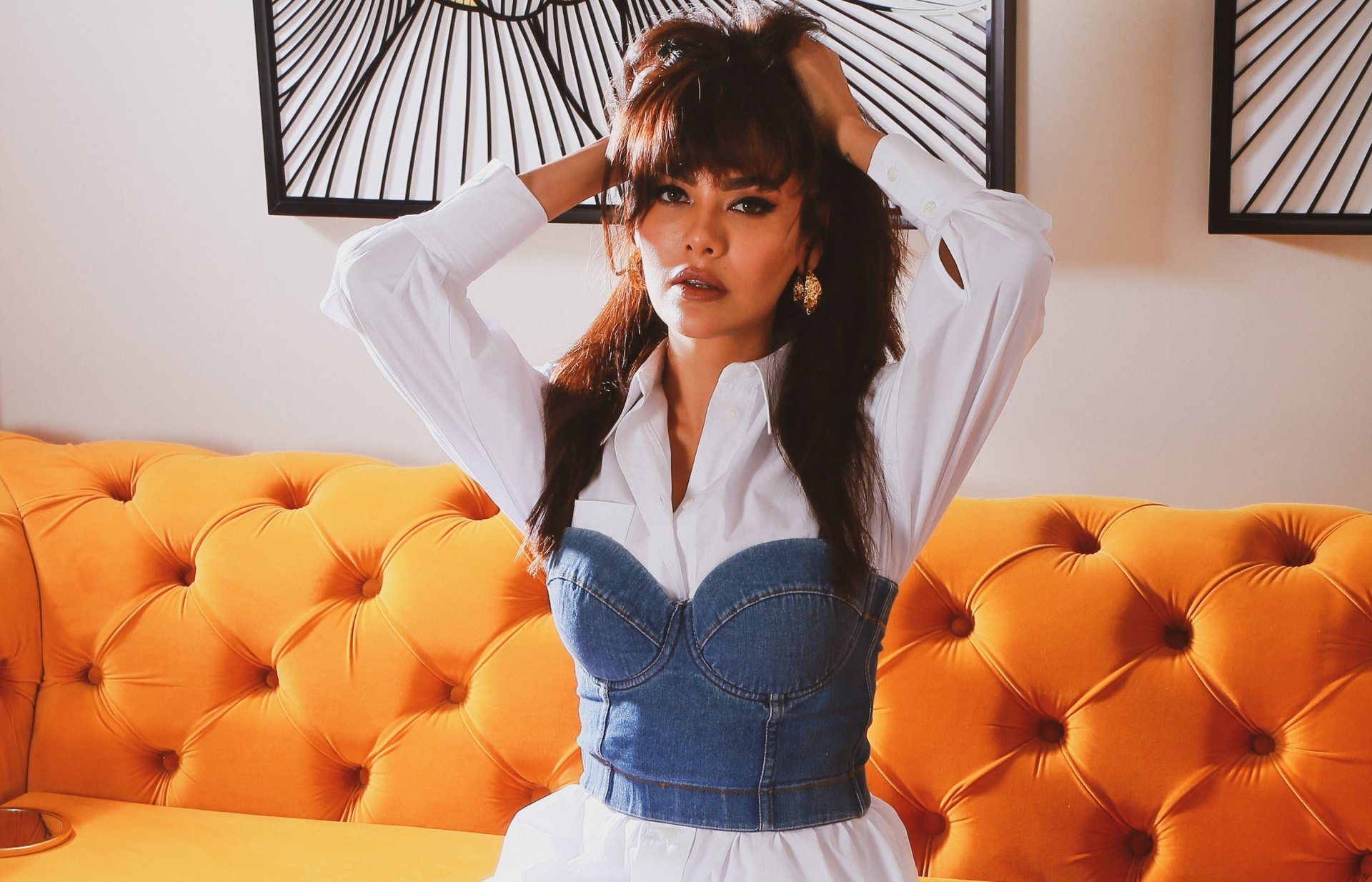 Female celebrity portrait on a bright orange tufted sofa, wearing a white shirt and denim corset, hands in her hair — 2K Quad HD PC desktop wallpaper/background.