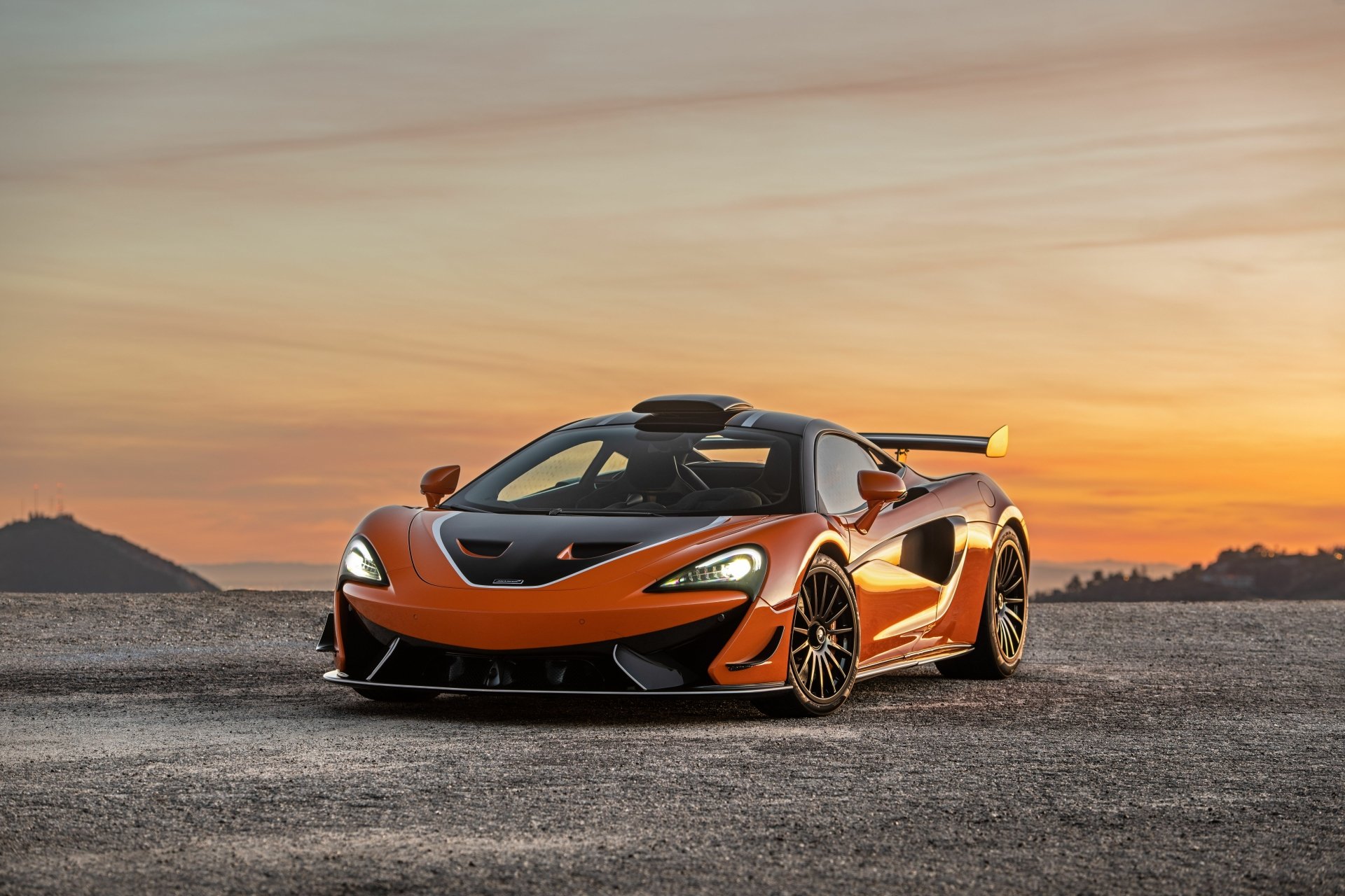 Orange McLaren 620R supercar parked on a textured ground with a scenic sunset background, captured in stunning 8K Ultra HD for PC desktop wallpaper.