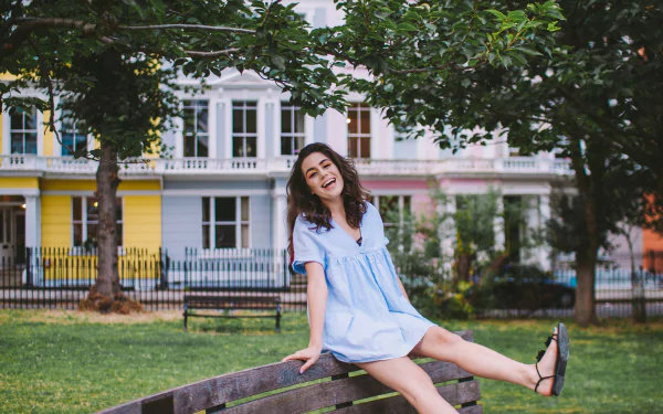 Happy woman in a blue dress sitting on a park bench with colorful houses in the background - HD dodie desktop wallpaper and background.
