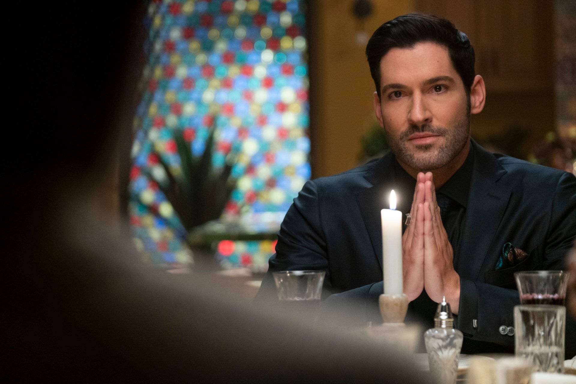 HD PC desktop wallpaper: Lucifer Morningstar (Lucifer TV show) seated at a candlelit table, hands clasped, with a colorful stained-glass window in the background.