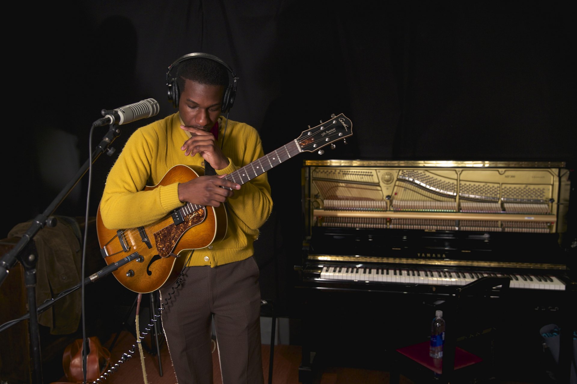 Musician in a yellow sweater playing guitar in a studio, with a grand piano in the background, suitable as an HD desktop wallpaper.