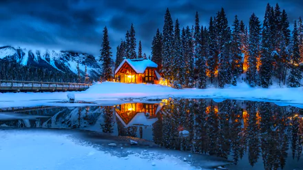 A cozy, snow-covered man-made cabin glows warmly beside a calm, reflective body of water, surrounded by tall pine trees under a deep blue winter sky.