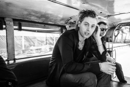 A black-and-white HD wallpaper featuring Luke Hemmings sitting inside a vehicle, casually engaged with his surroundings, capturing a moment of music and camaraderie.