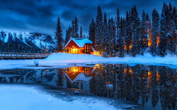 A cozy, snow-covered man-made cabin glows warmly beside a calm, reflective body of water, surrounded by tall pine trees under a deep blue winter sky.
