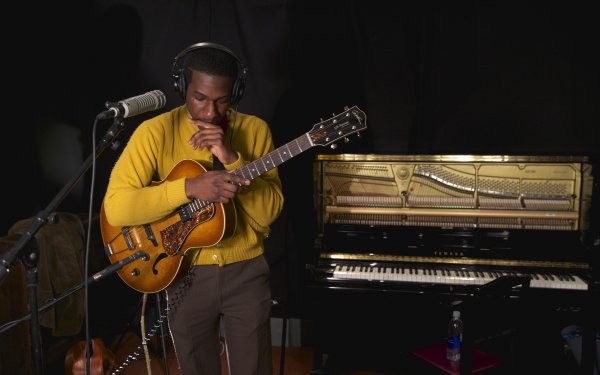 Musician in a yellow sweater playing guitar in a studio, with a grand piano in the background, suitable as an HD desktop wallpaper.