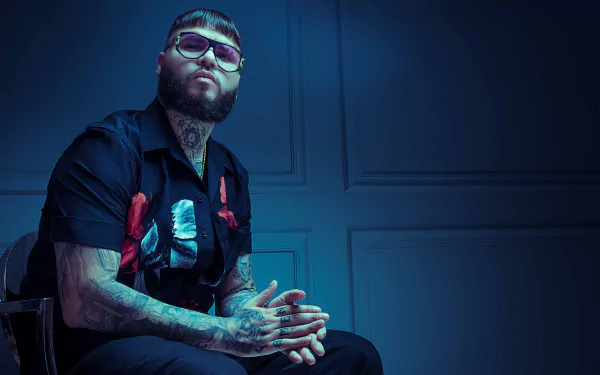 Stylish individual wearing sunglasses and a black shirt with red floral patterns, sitting confidently against a blue backdrop, featured in an HD desktop wallpaper and background.