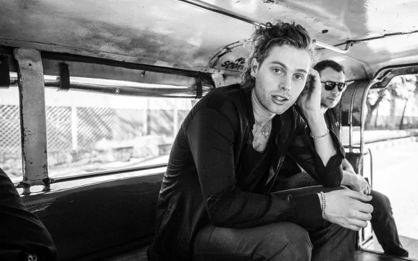 A black-and-white HD wallpaper featuring Luke Hemmings sitting inside a vehicle, casually engaged with his surroundings, capturing a moment of music and camaraderie.