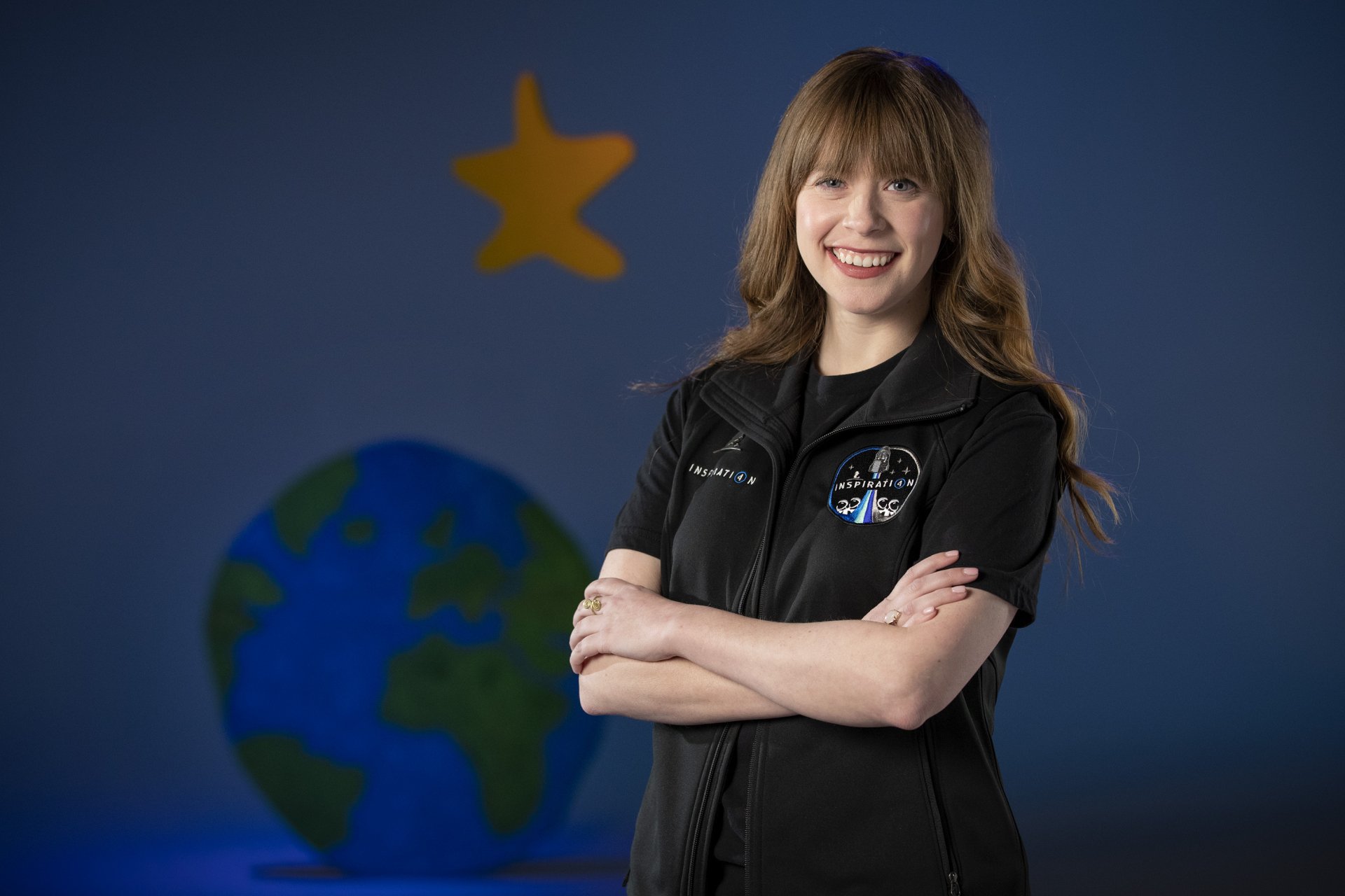 A confident woman poses with her arms crossed, wearing an astronaut-themed outfit, against a backdrop featuring a globe and a star, inspired by the Inspiration4 mission.