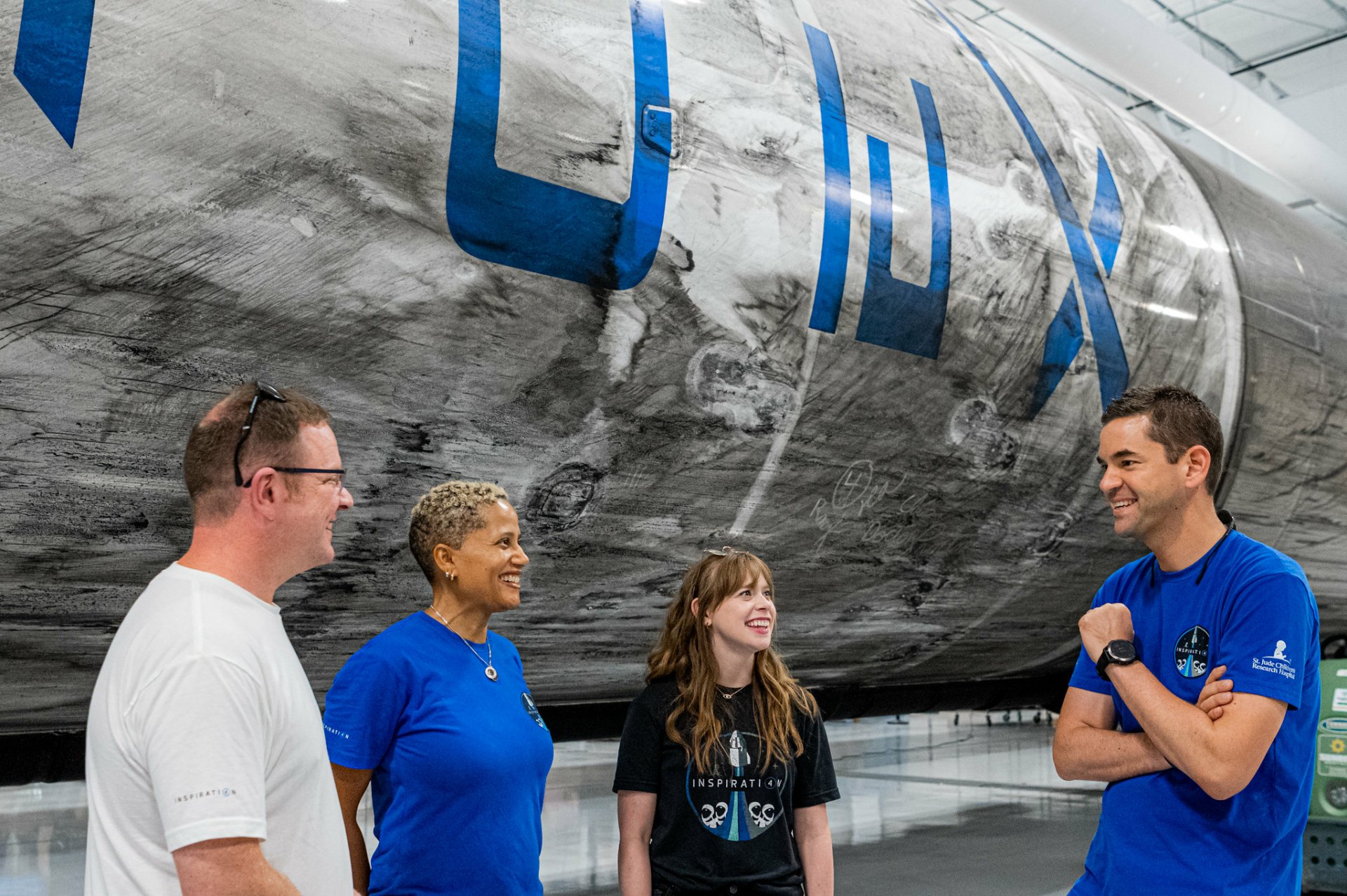 A group of four people engaged in conversation in front of a large rocket marked with Inspiration4, set in a bright, modern exhibition space. An inspiring scene reflecting innovation and exploration.