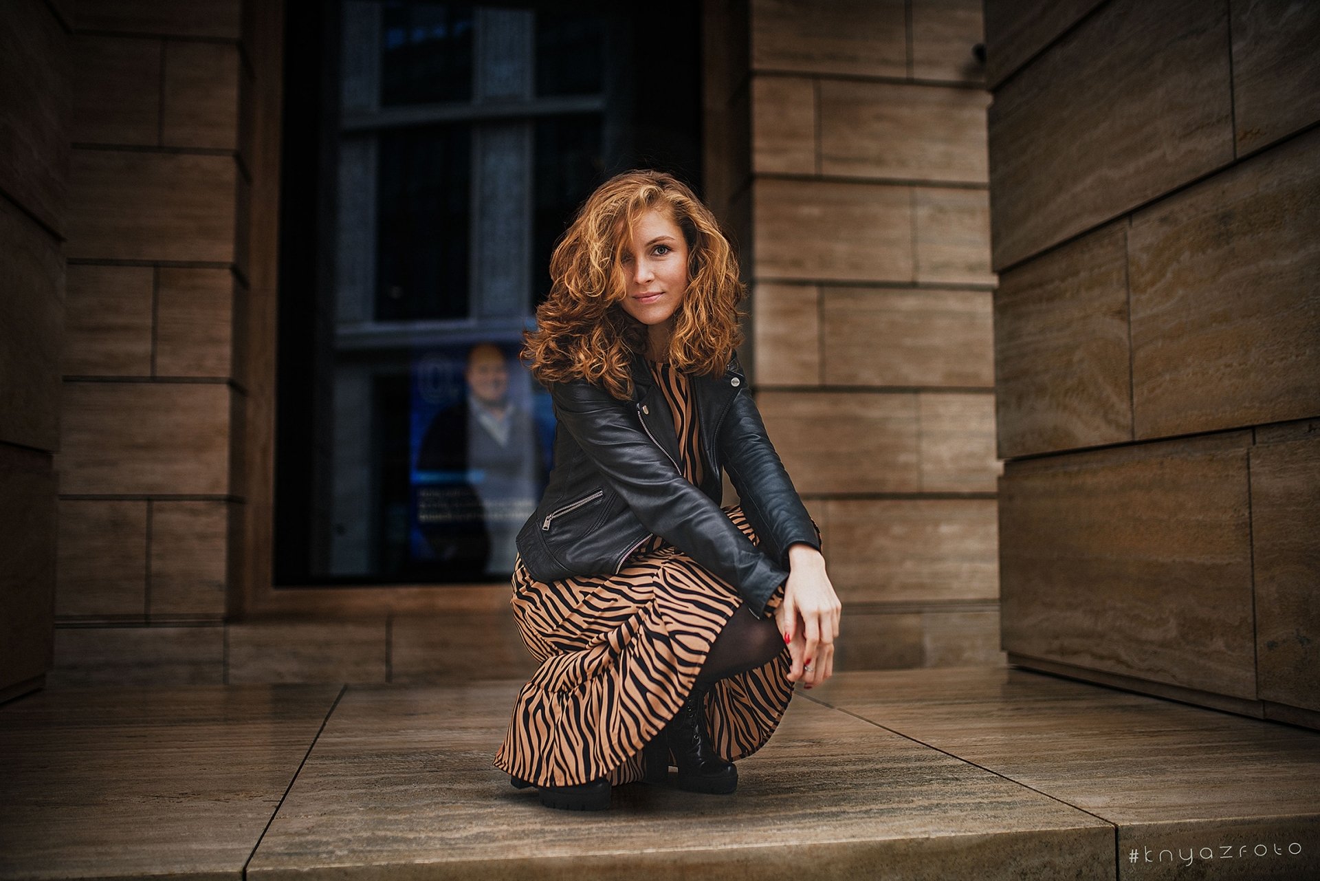 HD PC desktop wallpaper of a redhead woman model crouching in a leather jacket and patterned skirt against a warm, stone-paneled urban backdrop.