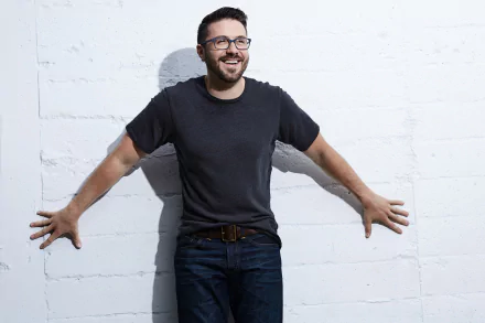 Smiling man in casual attire with glasses standing against a white brick wall for a HD desktop wallpaper and background.