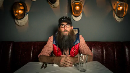 A bearded man wearing glasses and a cap, sitting at a table against a backdrop of stylish wall lamps, in an HD desktop wallpaper tagged with Crowder.