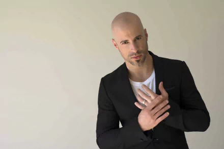 HD desktop wallpaper featuring a poised man with a bald head, wearing a black blazer over a white shirt, hand gently resting on his chest against a neutral backdrop.
