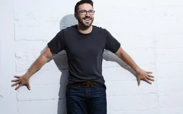 Smiling man in casual attire with glasses standing against a white brick wall for a HD desktop wallpaper and background.