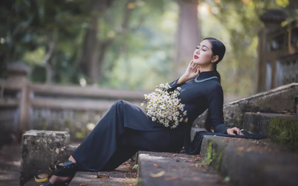 Moody 4K Ultra HD desktop wallpaper: a woman model in a flowing black dress reclines on mossy stone steps, eyes closed, holding a bouquet of small white flowers.