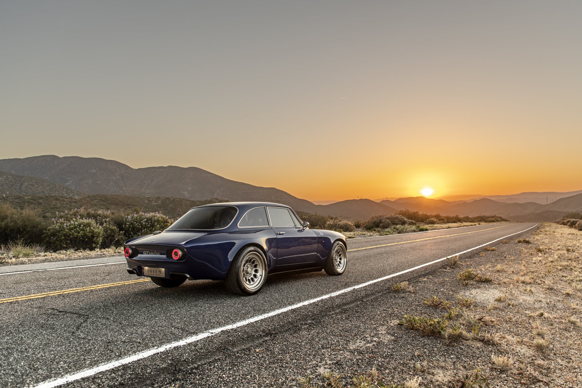 Totem Automobili GT Electric vehicle, an electric car on an open highway at sunset with mountains, presented as an 8K Ultra HD PC desktop wallpaper and background.