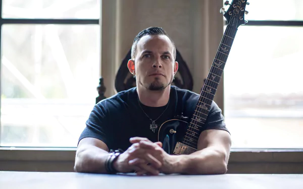Guitarist sitting at a table with a guitar beside him, gazing thoughtfully, in a room with window light - Tremonti themed HD desktop wallpaper and background.