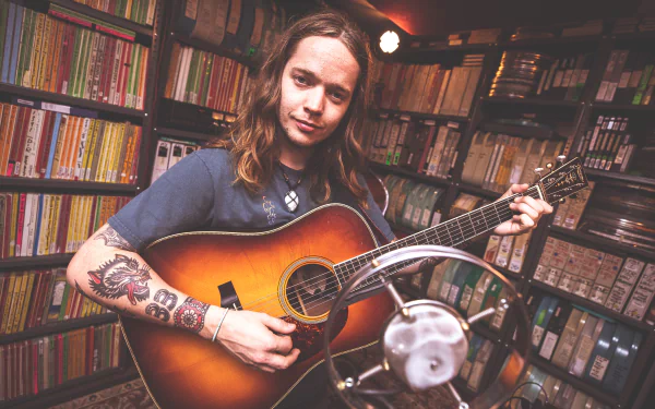 HD desktop wallpaper of a musician holding an acoustic guitar, set against a backdrop of bookshelves, tagged with Billy Strings.