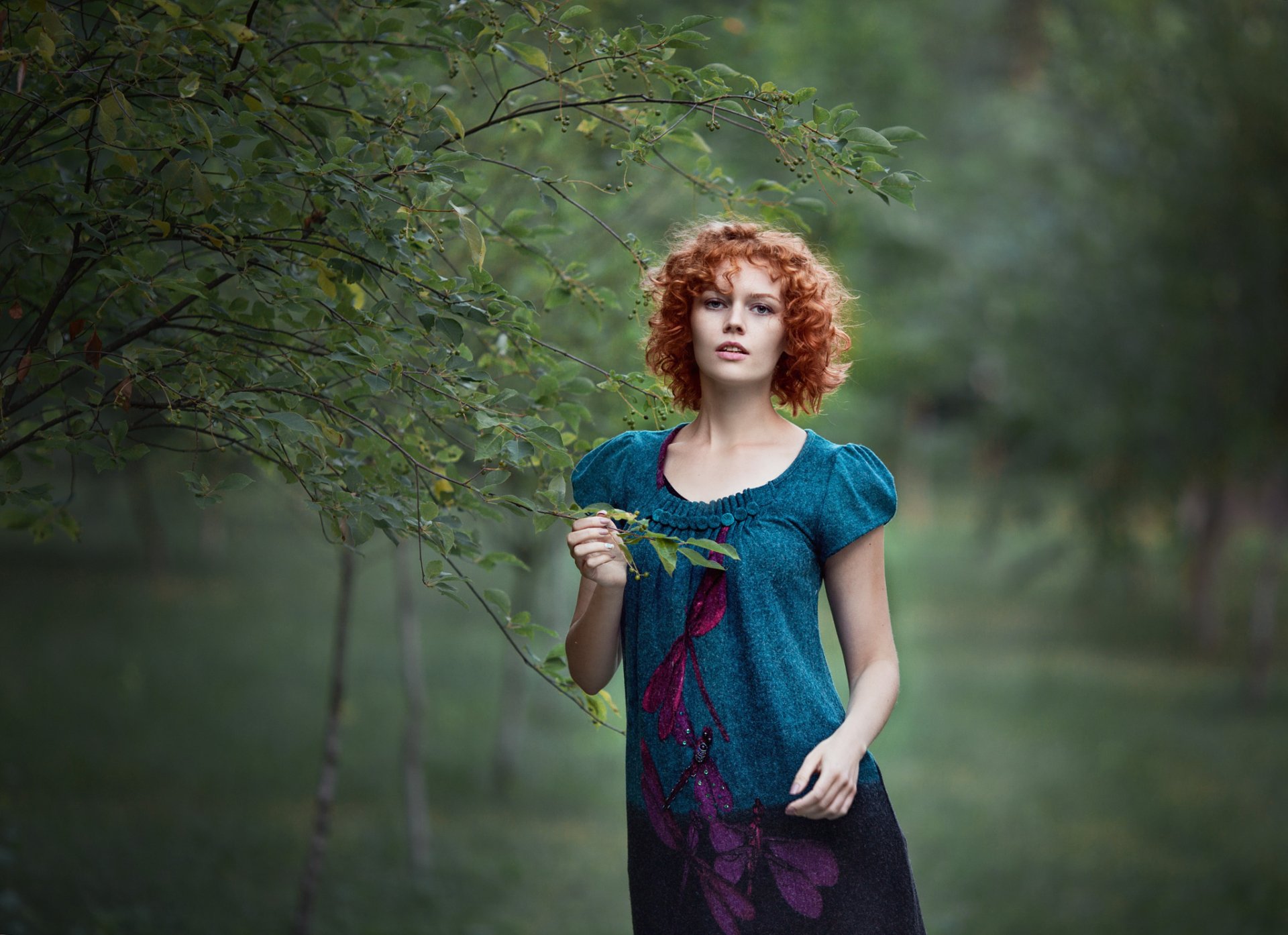 HD desktop wallpaper showing a redhead woman model in a teal dress standing amid lush green trees in a serene outdoor setting.