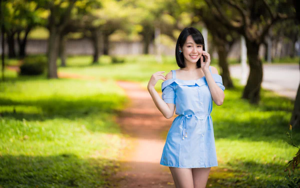 A smiling Asian woman in a blue dress stands on a sunlit park path with trees blurred in the background, captured in sharp 4K Ultra HD detail.