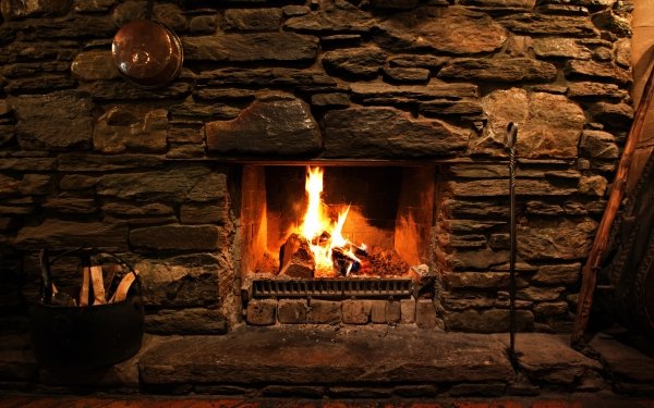 Alt Text: Cozy stone fireplace with roaring fire, fire irons, and basket of logs - warm HD desktop wallpaper and background.