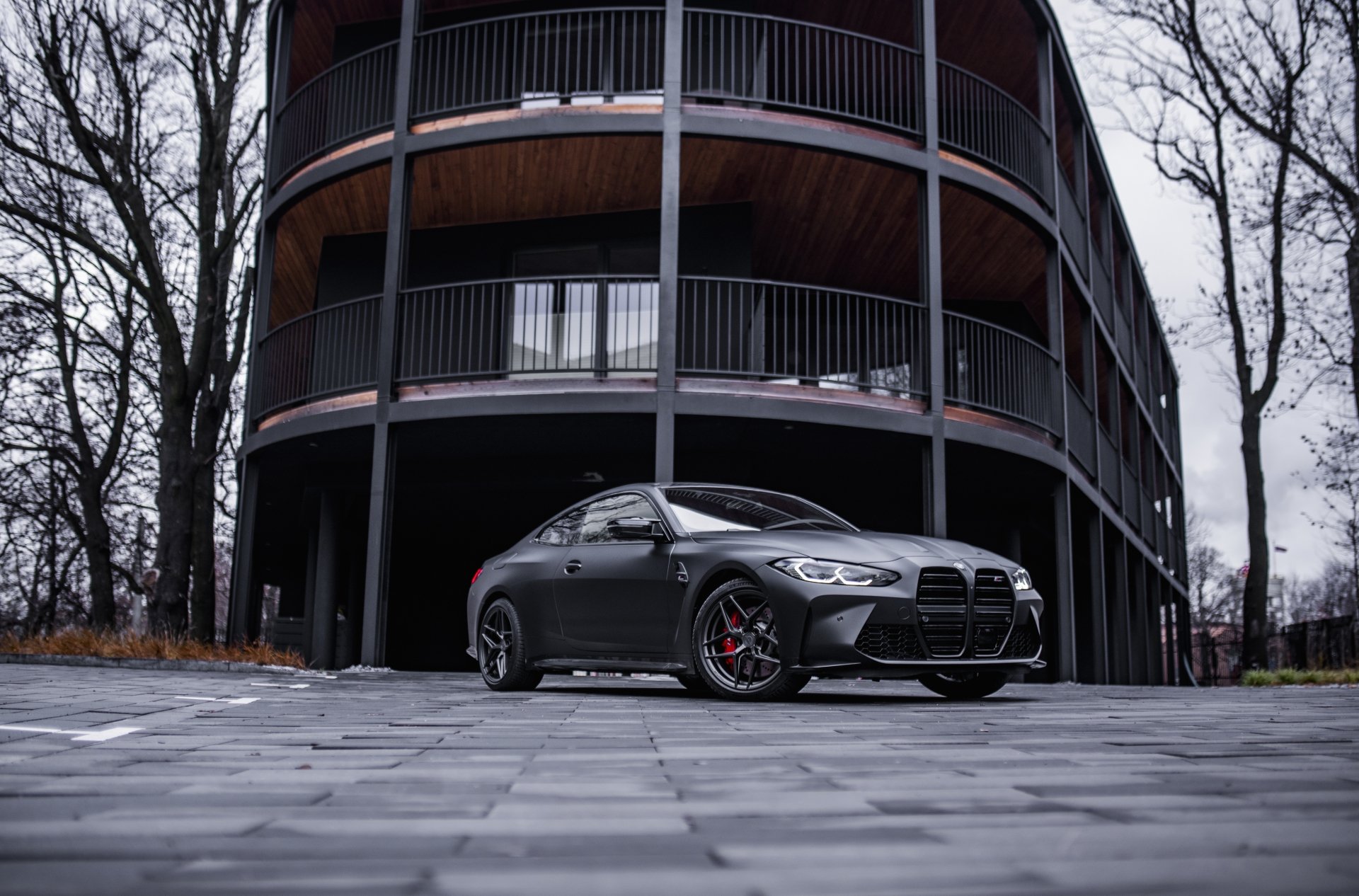 A sleek black BMW M4 parked on a paved area in front of a modern wooden balcony building, captured in 4K Ultra HD as a PC desktop wallpaper.