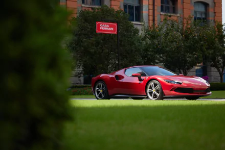 Red Ferrari 296 GTB supercar parked on lawn before an urban building, captured as an 8K Ultra HD PC desktop wallpaper/background.
