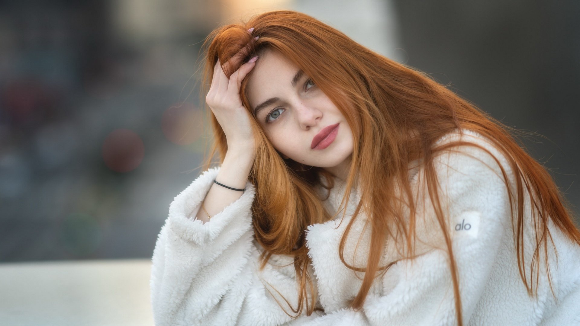 HD desktop wallpaper featuring a redhead woman model with long vibrant hair, wearing a white fuzzy jacket, posing with her hand in her hair against a blurred background.