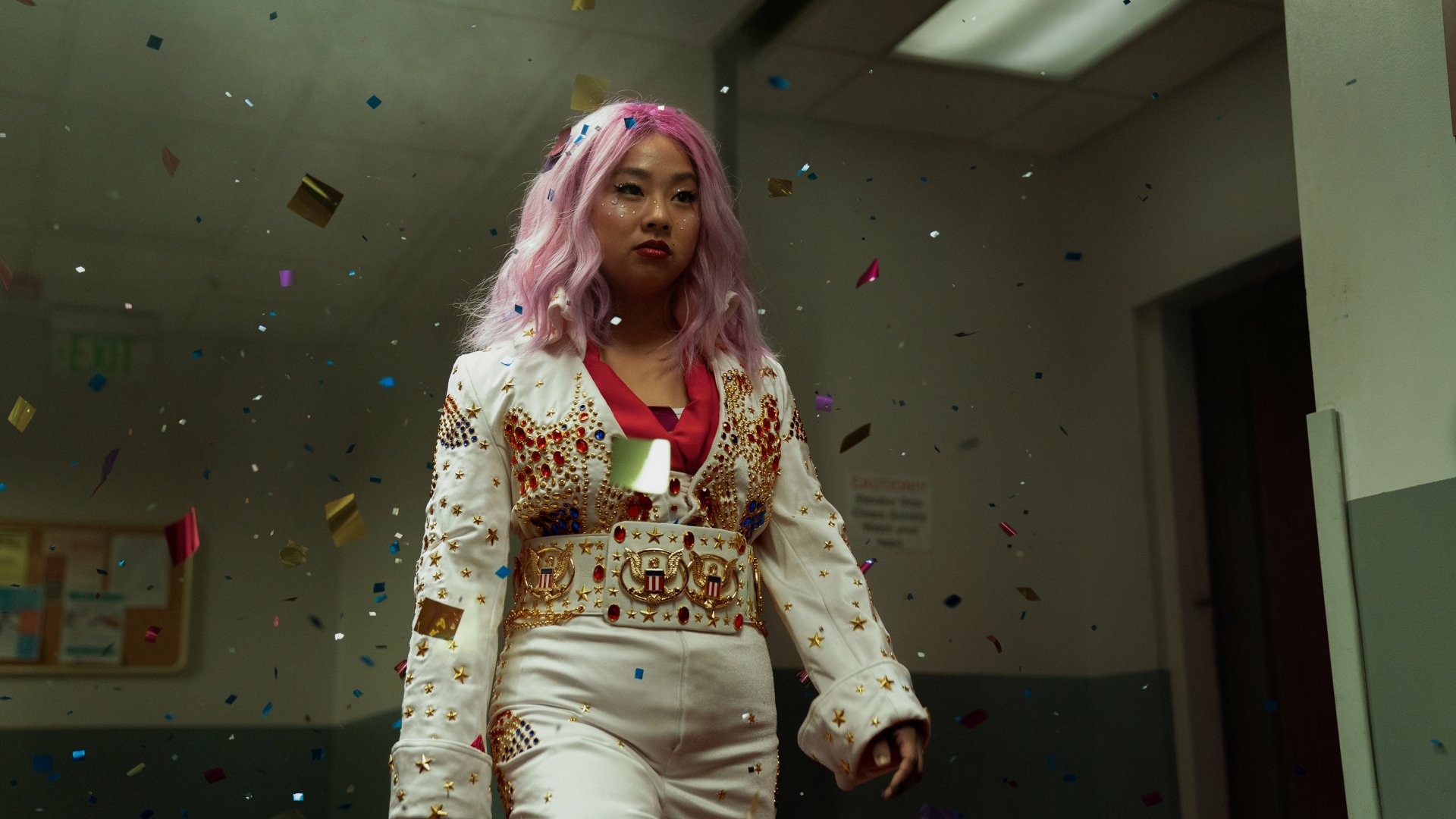 2K Quad HD desktop wallpaper: woman with pink hair in embellished white jumpsuit strides through a dim hallway as confetti rains down — cinematic, surreal movie still.