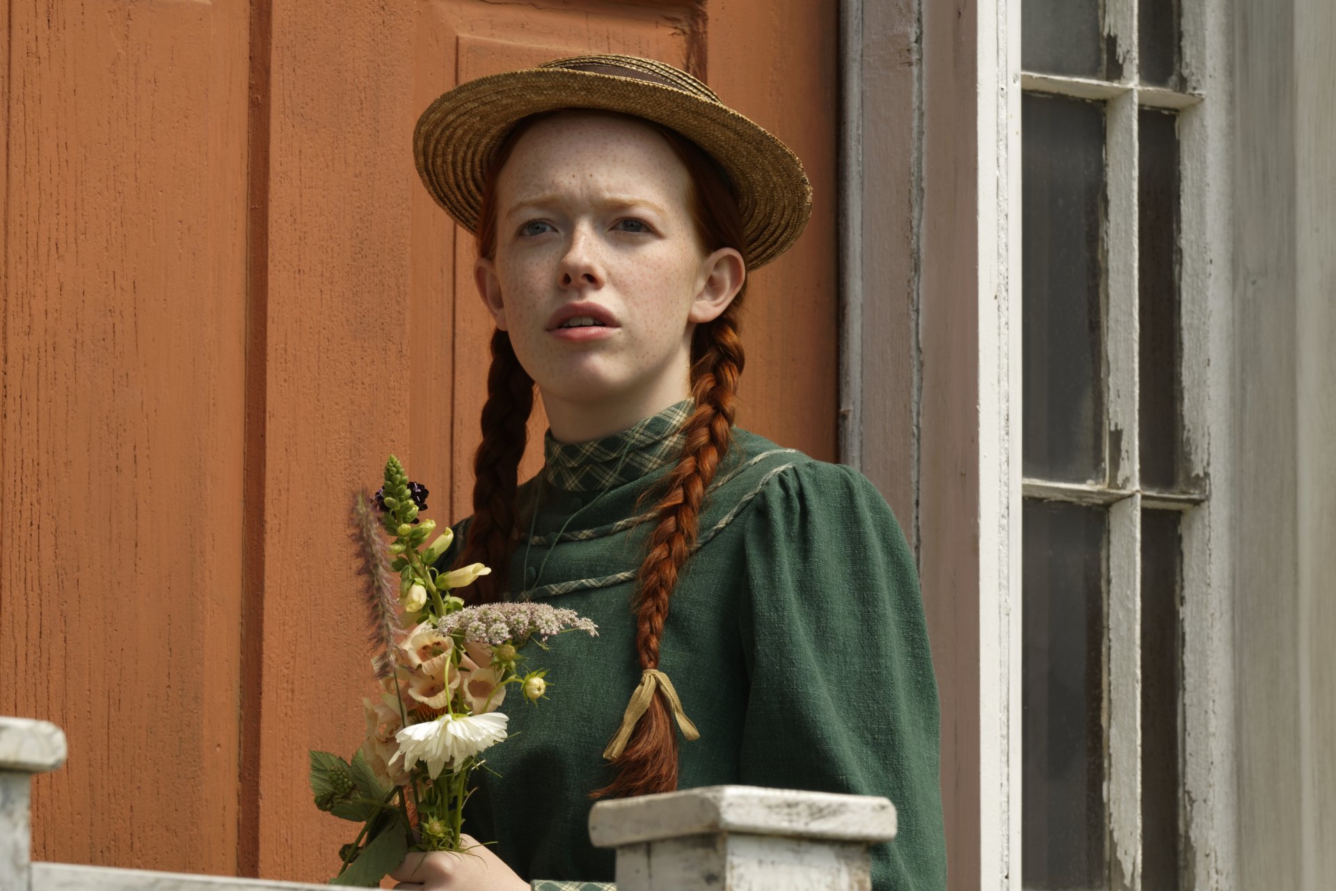 2K Quad HD PC desktop wallpaper: red‑haired girl in braids and green period dress holding flowers on a porch — scene from the TV show Anne with an E.