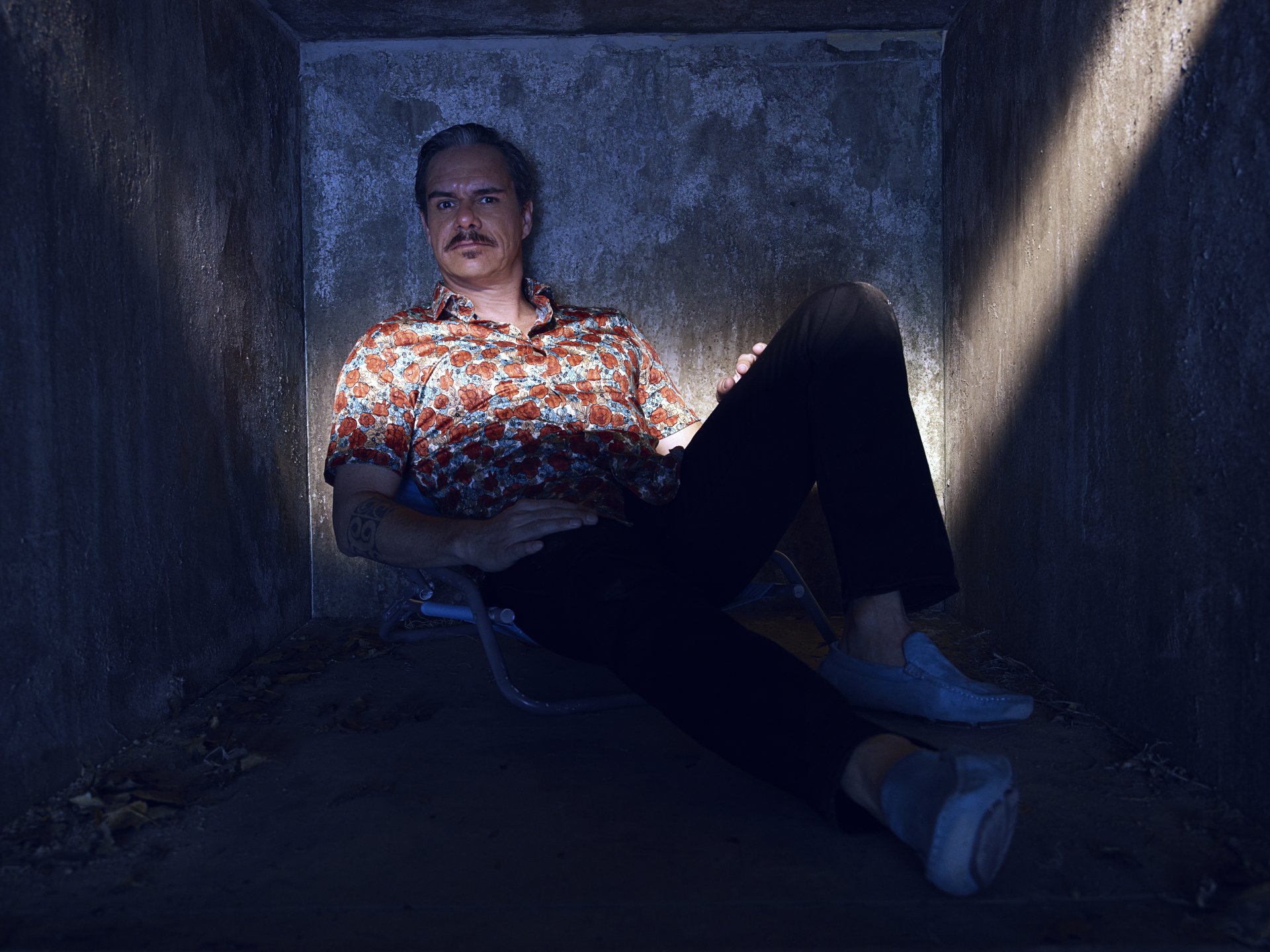 4K Ultra HD desktop wallpaper of Lalo Salamanca from Better Call Saul, reclining in a dim concrete shaft, patterned shirt catching a narrow beam of sunlight.
