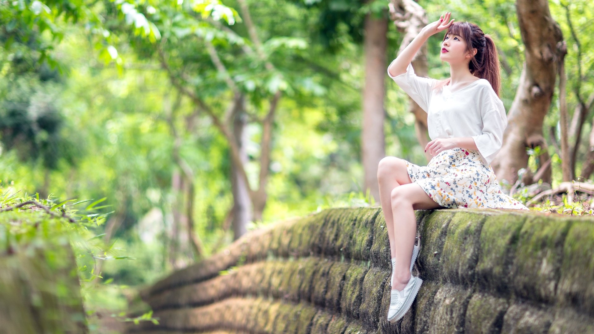 Asian woman in casual outfit sitting on a mossy stone wall surrounded by lush greenery, captured in vibrant detail for a 4K Ultra HD PC desktop wallpaper.