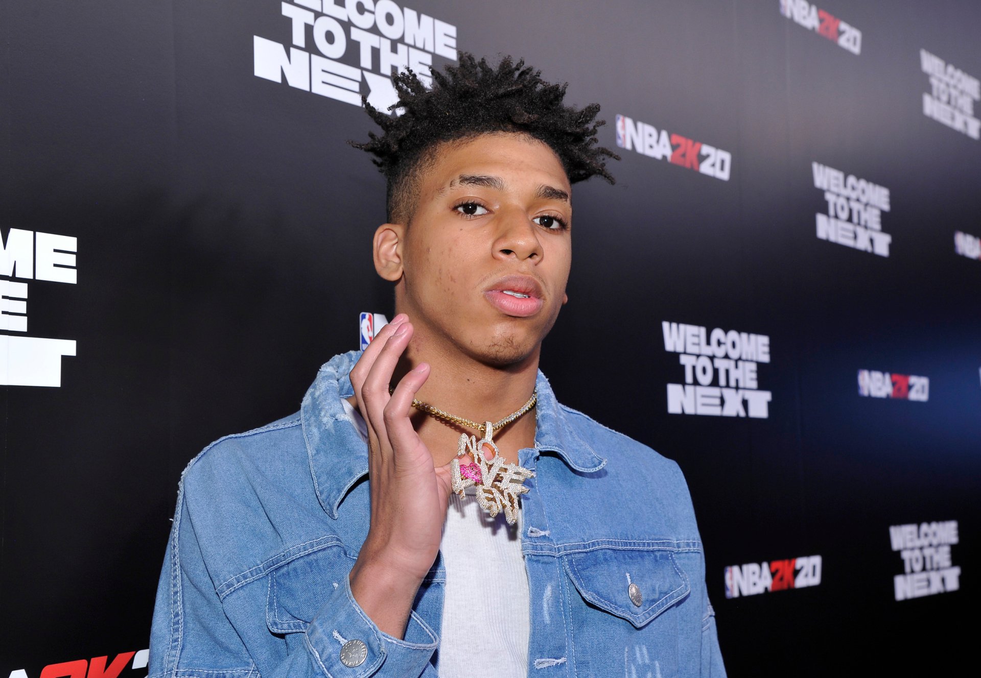 2K Quad HD PC desktop wallpaper of a young hip-hop artist at a music red carpet, denim jacket and necklace, posed against a dark branded backdrop.
