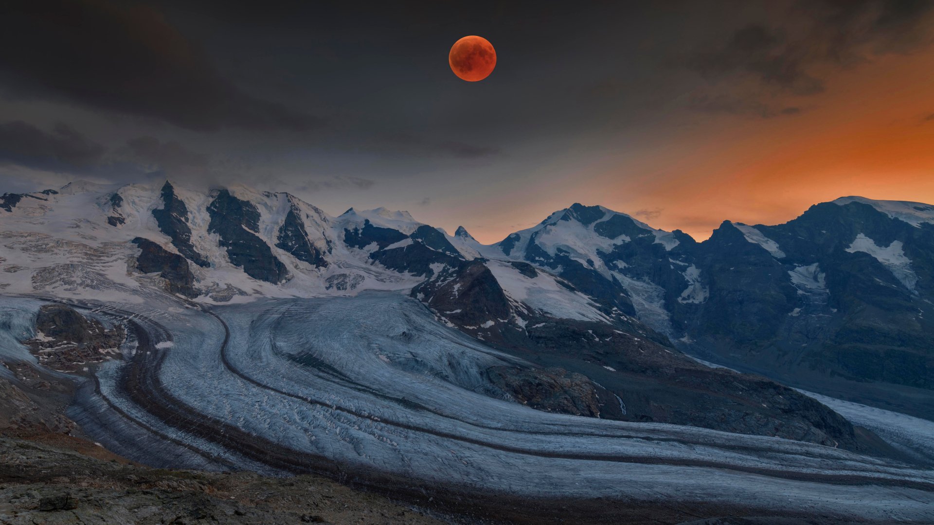 4K Ultra HD image of a blood moon lunar eclipse over the snow-covered Alps, highlighting rugged peaks and a winding glacier under a darkening sky.