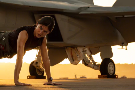 Monica Barbaro from Top Gun: Maverick performing push-ups on the tarmac near a jet, captured in an HD movie wallpaper background.